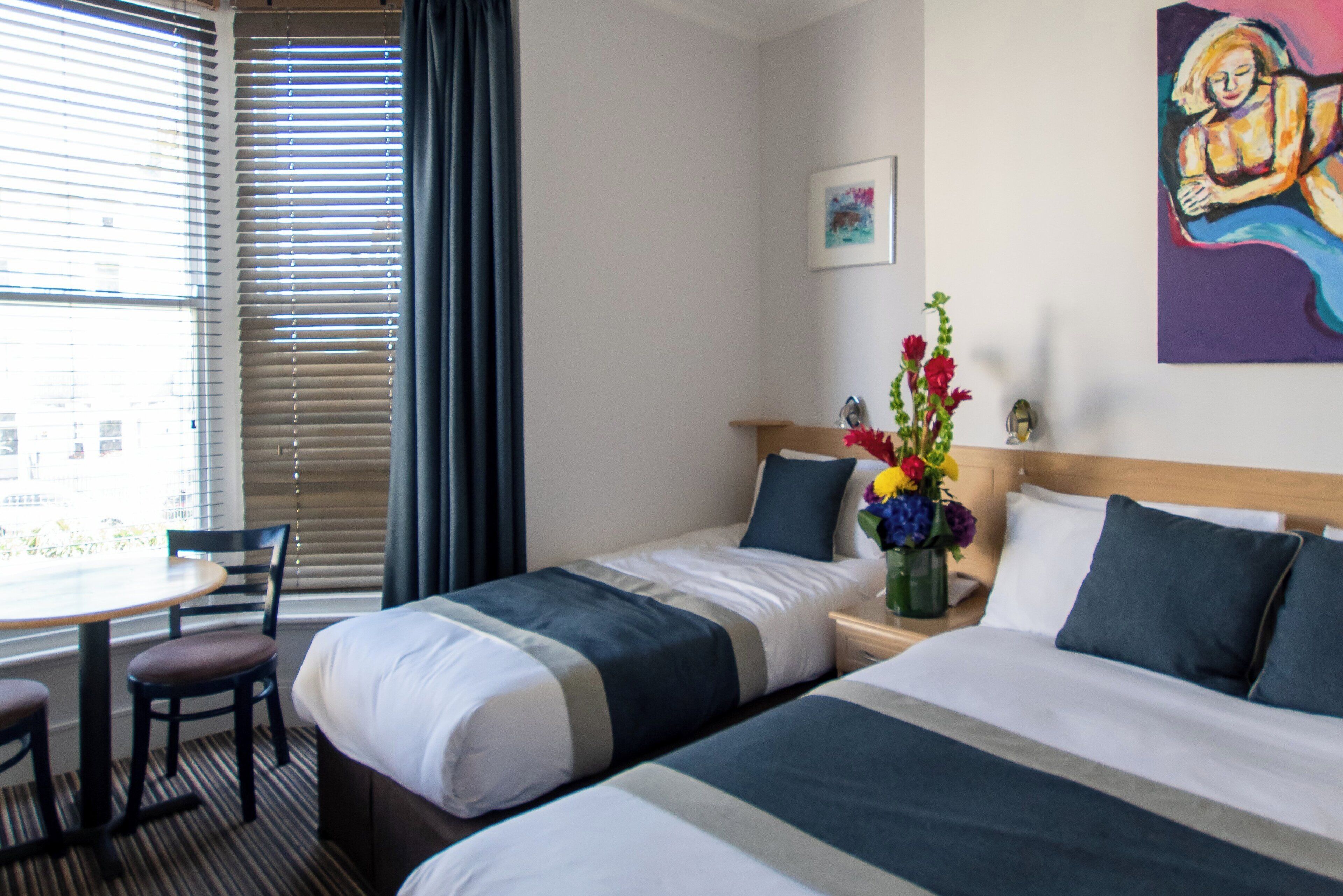 A hotel room with two beds, a dining table with chairs, and a window with blinds.