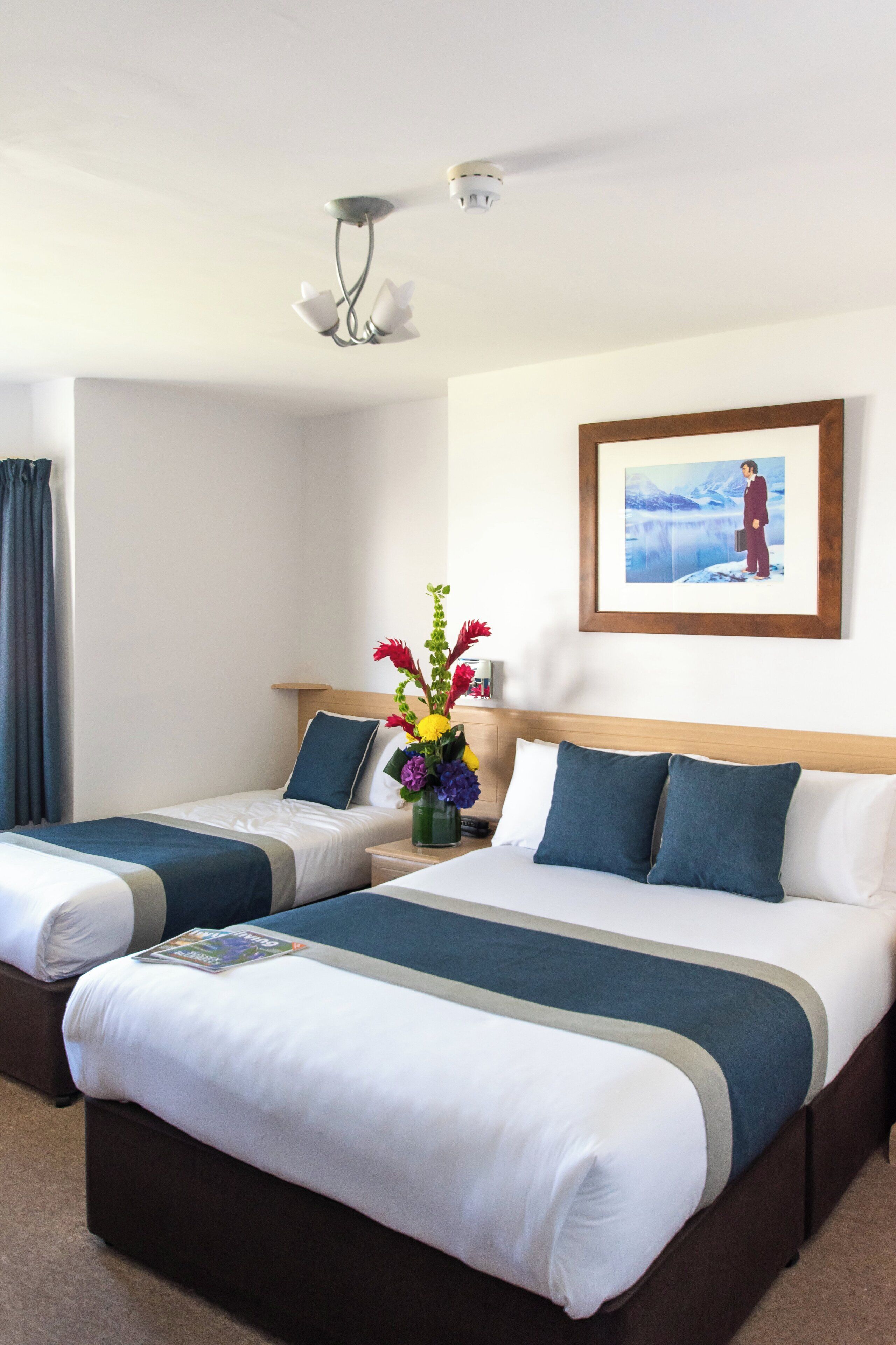 Ein Hotelzimmer mit zwei Betten, einem Nachttisch mit Blumenvase, einem gerahmten Bild an der Wand und einer an der Decke befestigten Überwachungskamera.