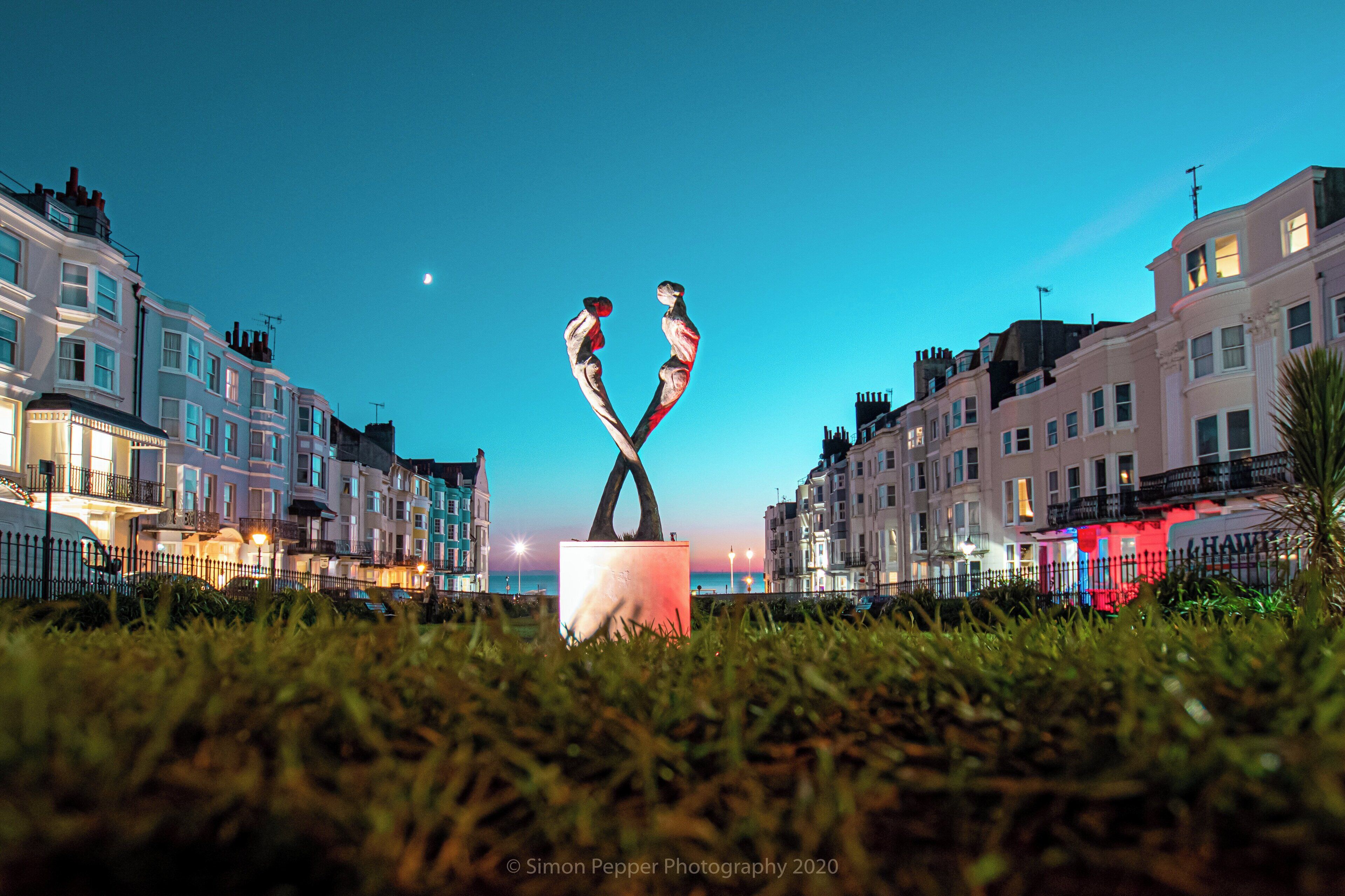 A sculpture of two figures in a garden setting with buildings in the background.
