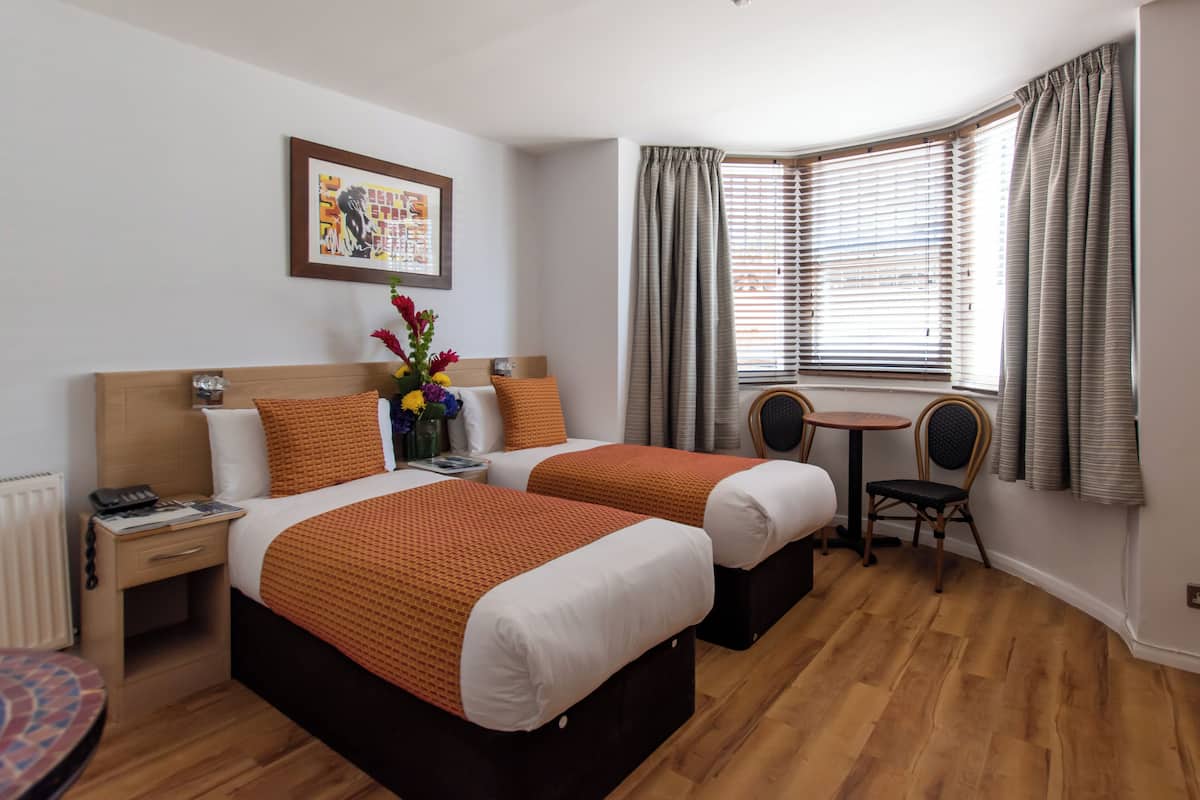 A hotel room with two beds, a small table, a chair, a picture on the wall, and a window with curtains.