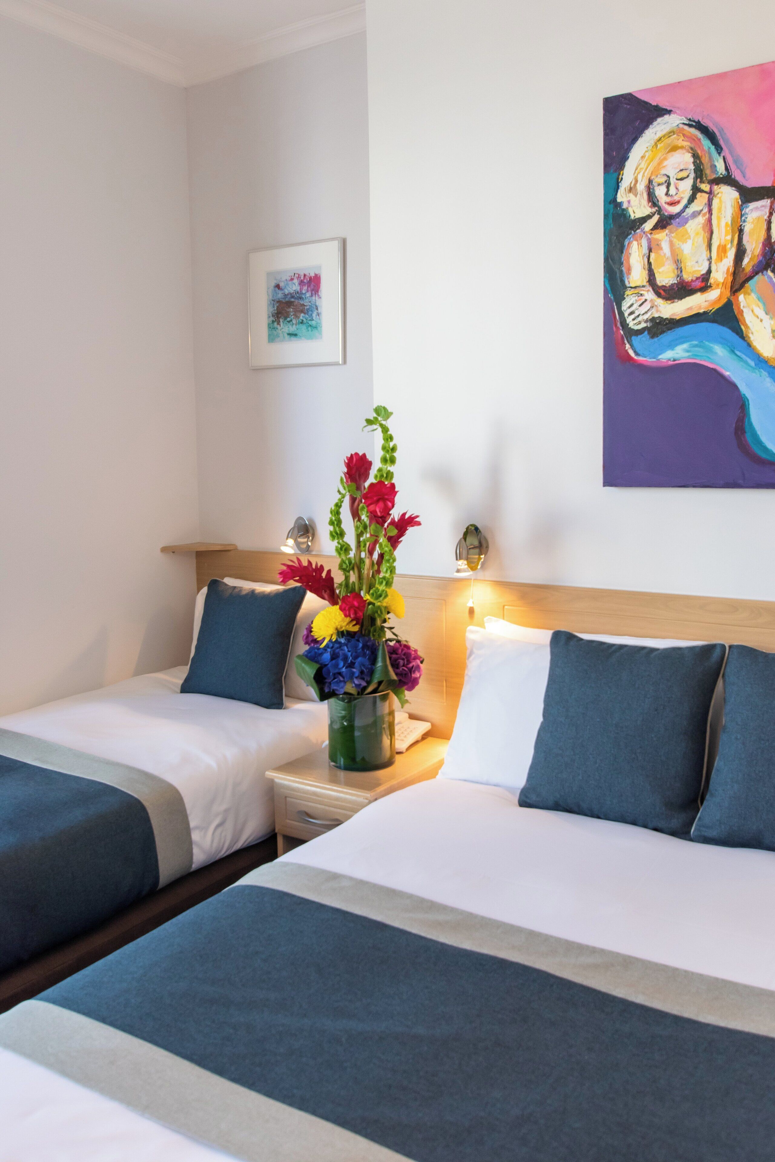 Une chambre d’hôtel avec deux lits, une table de chevet, un vase de fleurs et un tableau au mur.