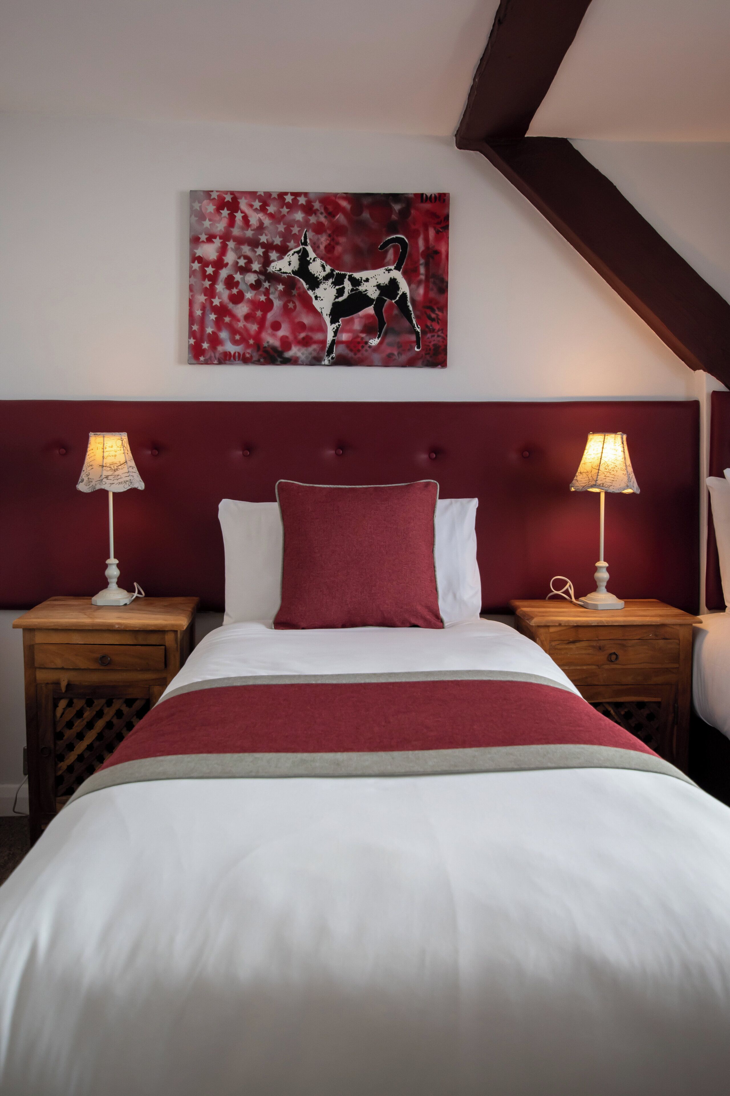 Ein ordentlich bezogenes Bett mit roter Tagesdecke, zwei Nachttischlampen, einem roten Kopfteil und einem Bild von einem Hund an der Wand.