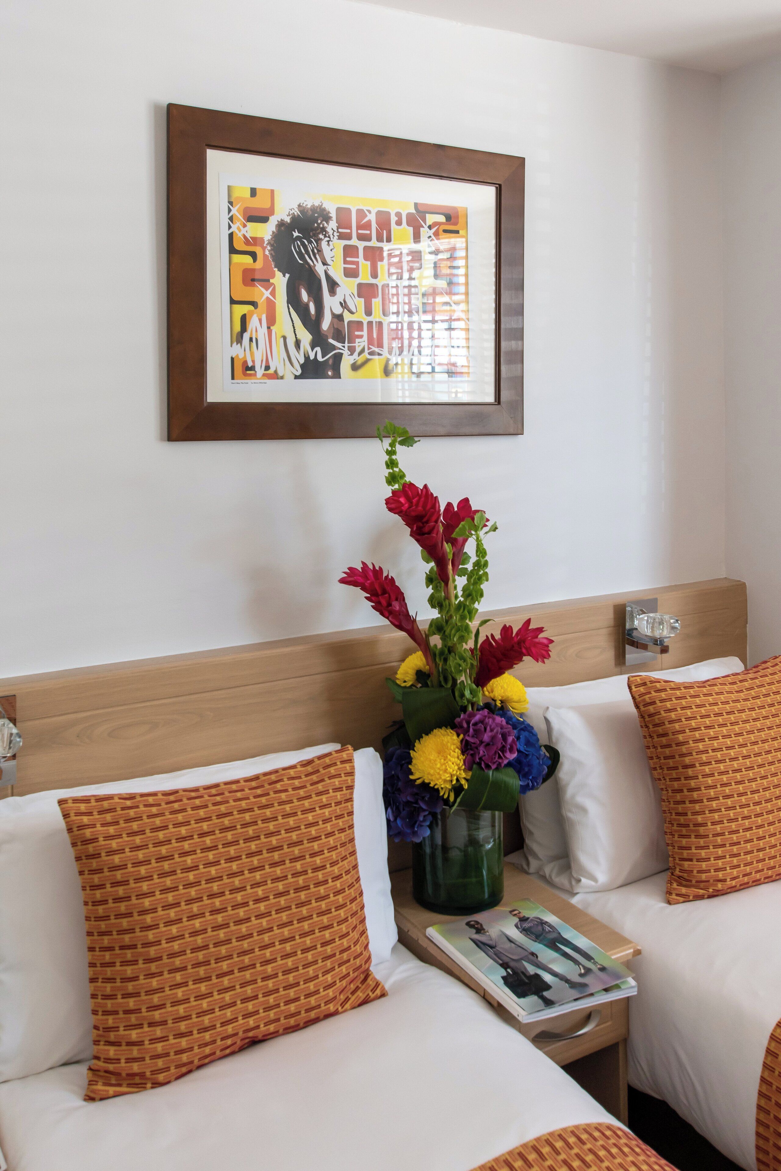 A hotel room with two beds, a framed artwork on the wall, a vase with flowers, and a magazine on the nightstand.