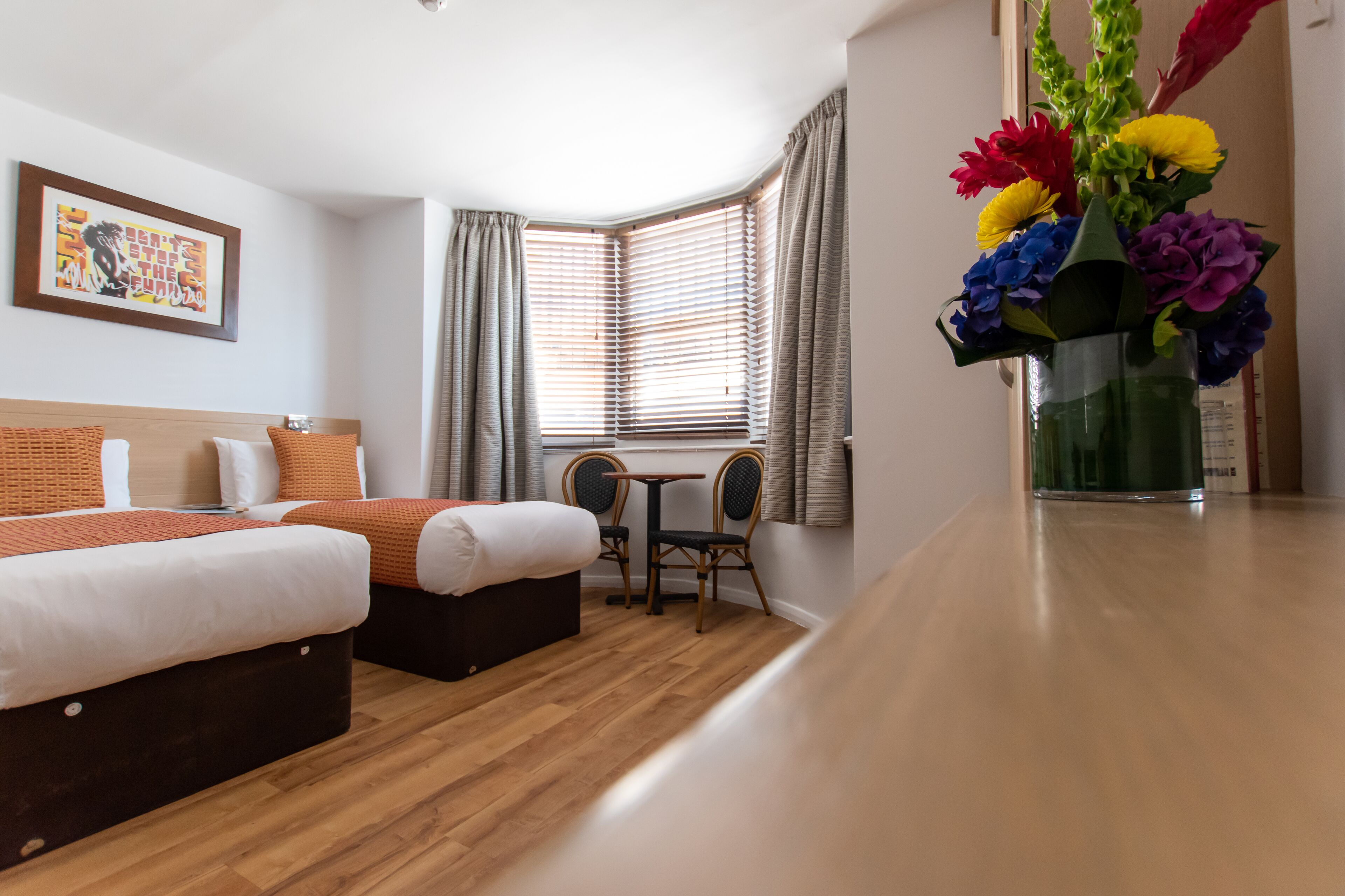 A hotel room with two beds, a small table with chairs, a window with blinds, and a vase with flowers.