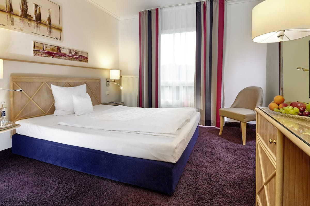 A hotel room with a bed, a chair, a desk with a lamp, a mirror, and a fruit basket.