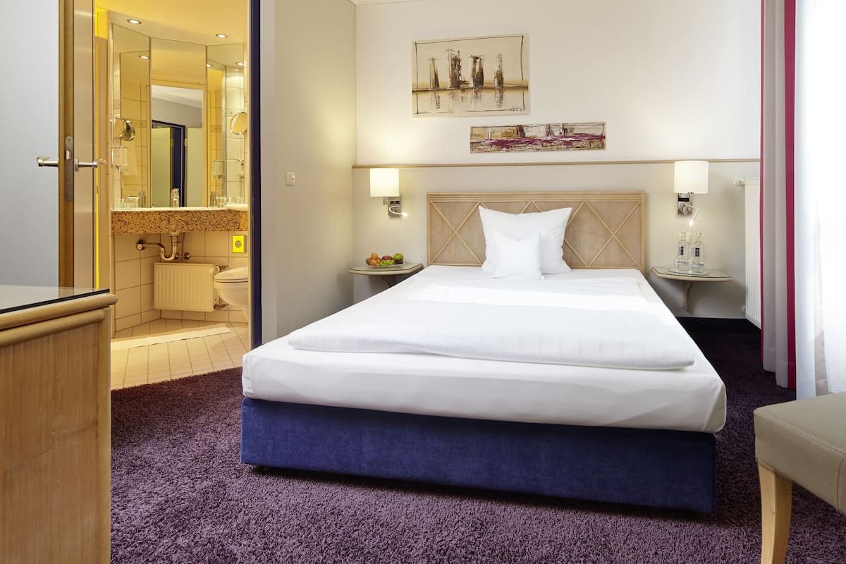 A hotel room with a large bed, a bedside table with a lamp, a small table with fruit, and a mirror in the bathroom.
