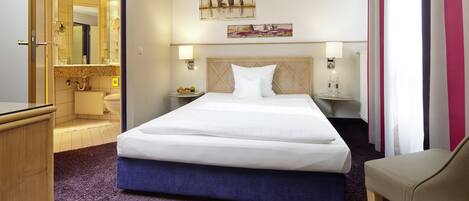 A hotel room with a large bed, a bedside table with a lamp, a small table with fruit, and a mirror in the bathroom.