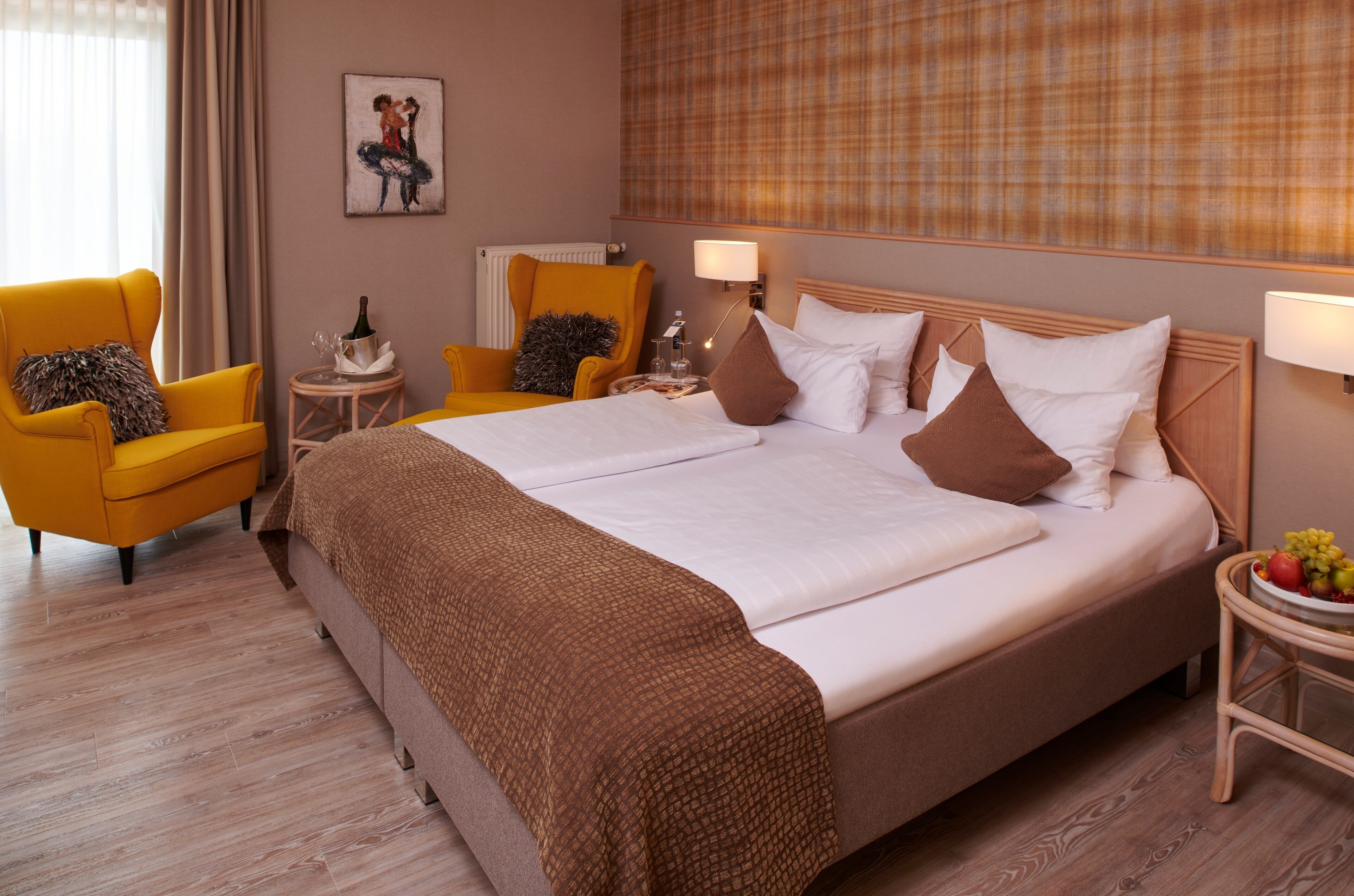 A hotel room with a bed, two yellow armchairs, a small round table with a lamp, and a fruit bowl on the side table.