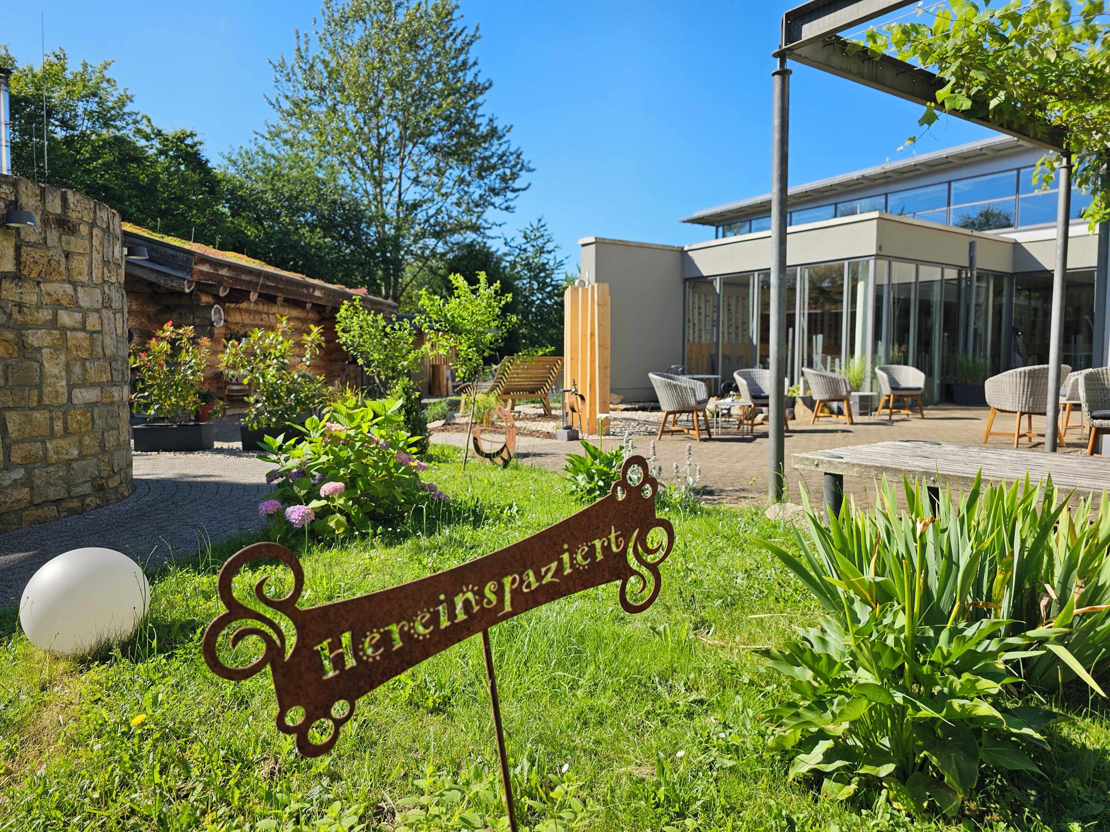 Ein Gartenbereich mit einem Metallschild, Sitzgelegenheiten im Freien und einem modernen Gebäude im Hintergrund.