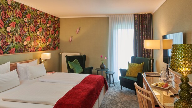 A hotel room with a large bed, a desk with a lamp, a chair, a small table, and a decorative vase. - Parkhotel Weiskirchen (Weiskirchen)