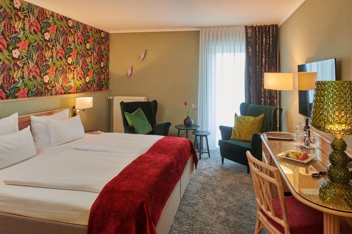 A hotel room with a large bed, a desk with a lamp, a chair, a small table, and a decorative vase.