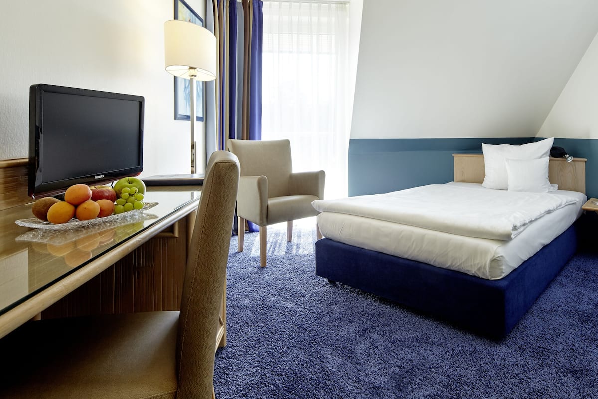 A hotel room with a bed, a desk with a television, a chair, and a nightstand with a lamp.