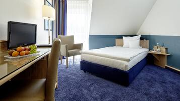 A hotel room with a bed, a desk with a television, a chair, and a nightstand with a lamp.