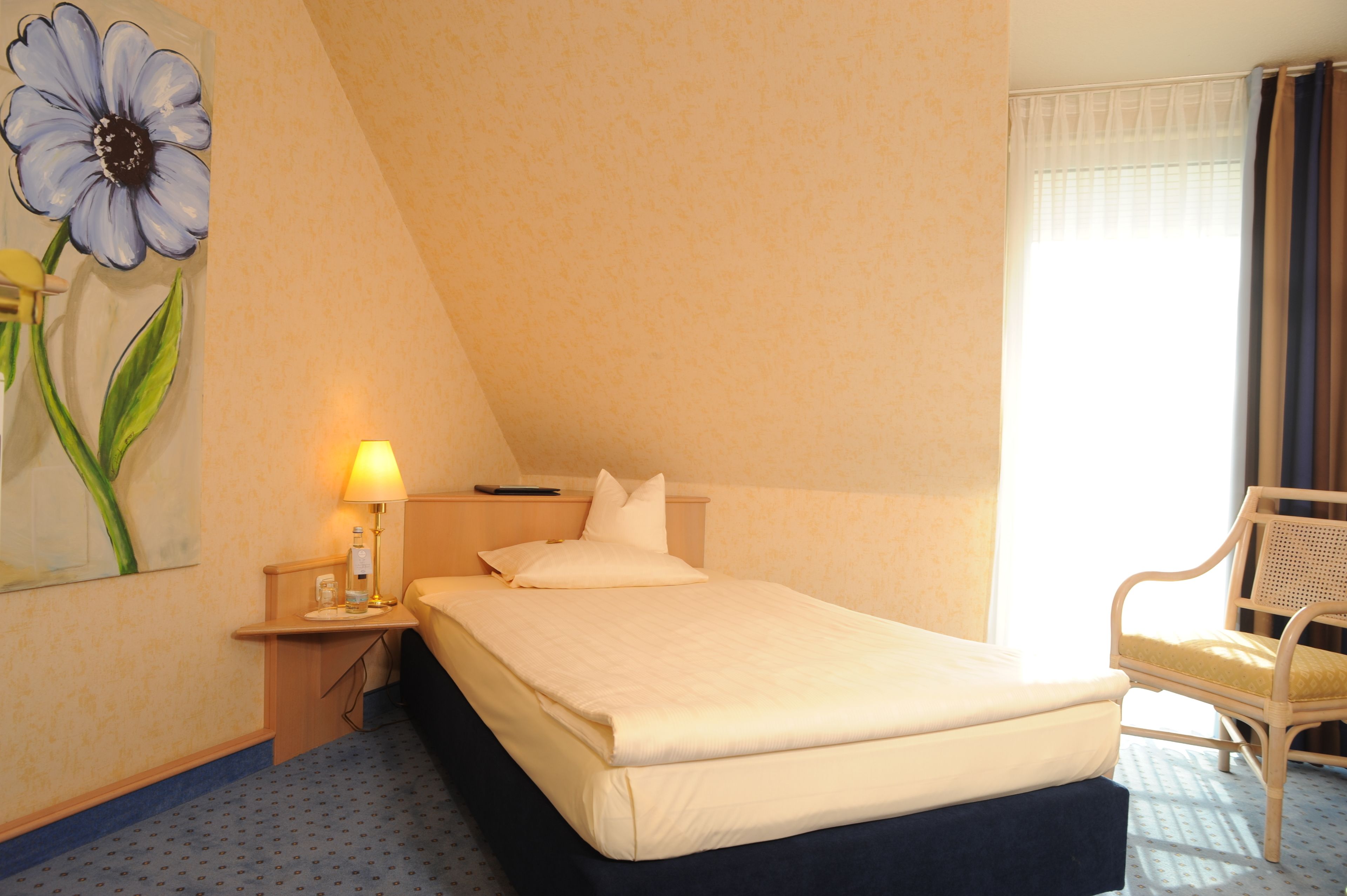 Hotellihuone, jossa on sänky, yöpöytä lampun kanssa, tuoli ja seinällä oleva kukka-aiheinen taulu.