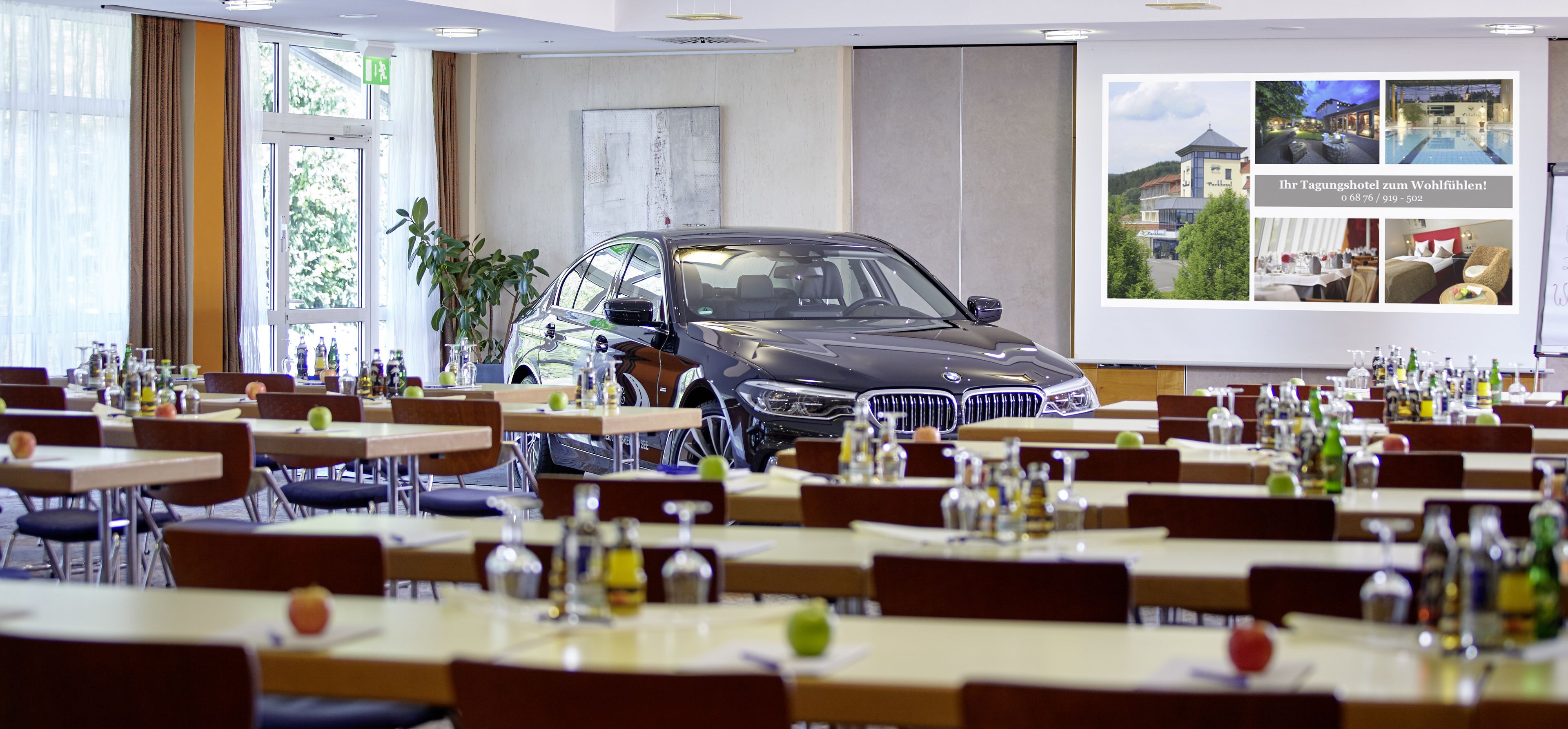 Un comedor con mesas dispuestas para comer, un coche BMW estacionado al fondo y un ventanal con cortinas.