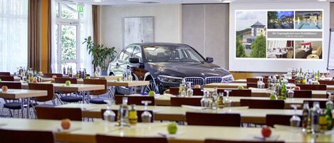 Un comedor con mesas dispuestas para comer, un coche BMW estacionado al fondo y un ventanal con cortinas.