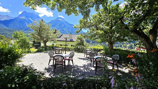 A patio with outdoor seating, a building with a sloped roof, and mountains in the background. - Typically Swiss Hotel Altana (Scuol)