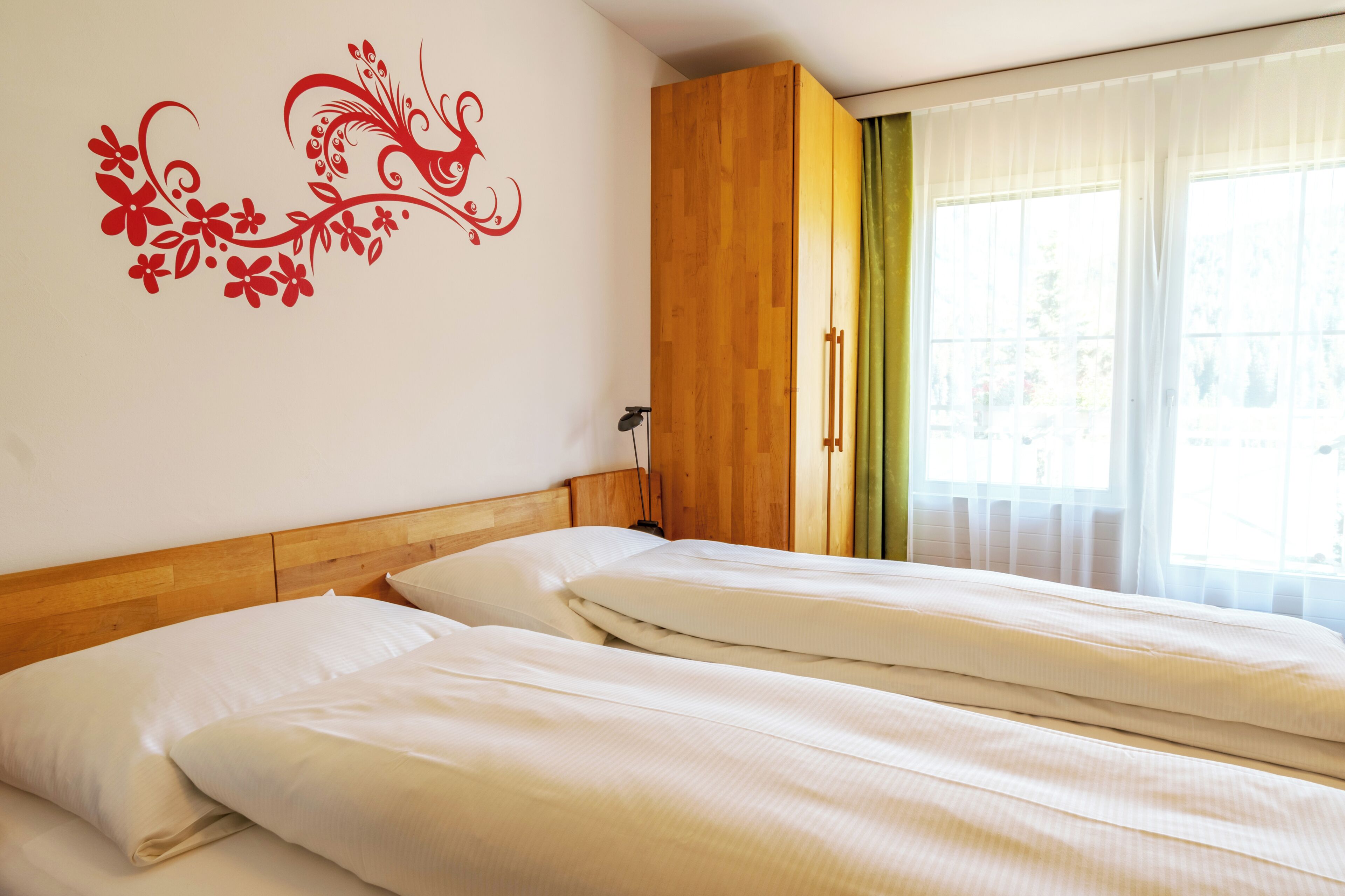 A hotel room with two beds, a wooden wardrobe, a decorative wall sticker, and a window with curtains.