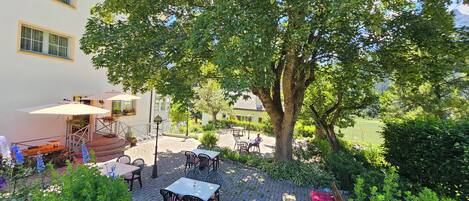 An outdoor patio with tables and chairs, surrounded by greenery and a large tree.