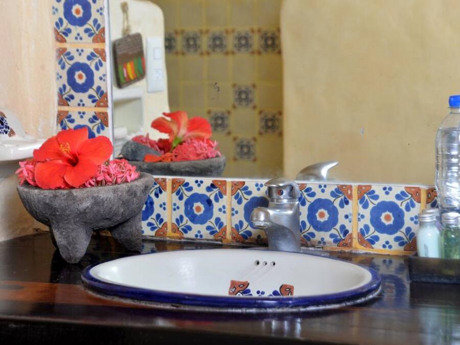 A bathroom with a sink, mirror, and decorative tiles. There are flower arrangements in a stone basin and bottles on the counter.