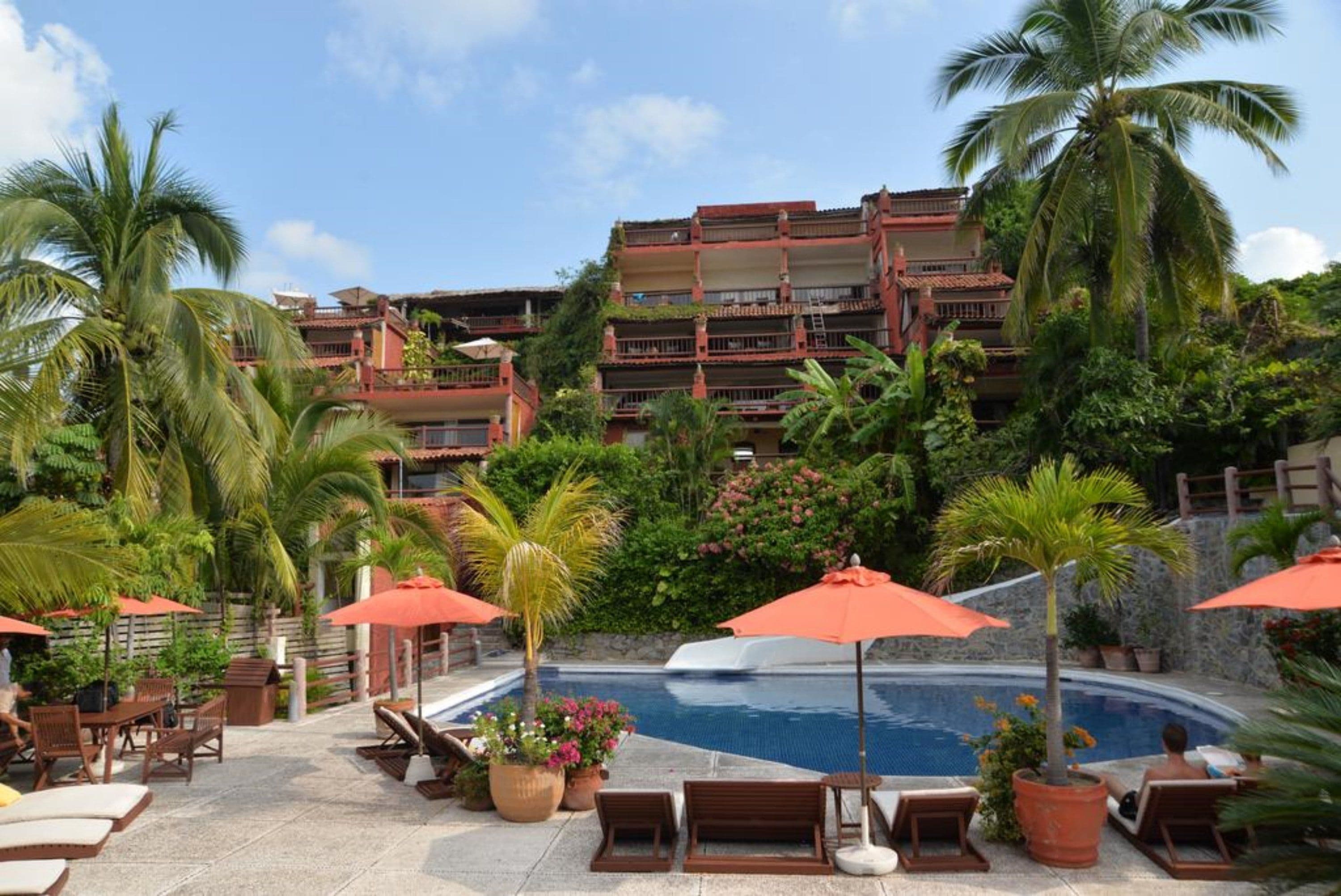 A pool area with lounge chairs and umbrellas, surrounded by palm trees and a building with balconies.