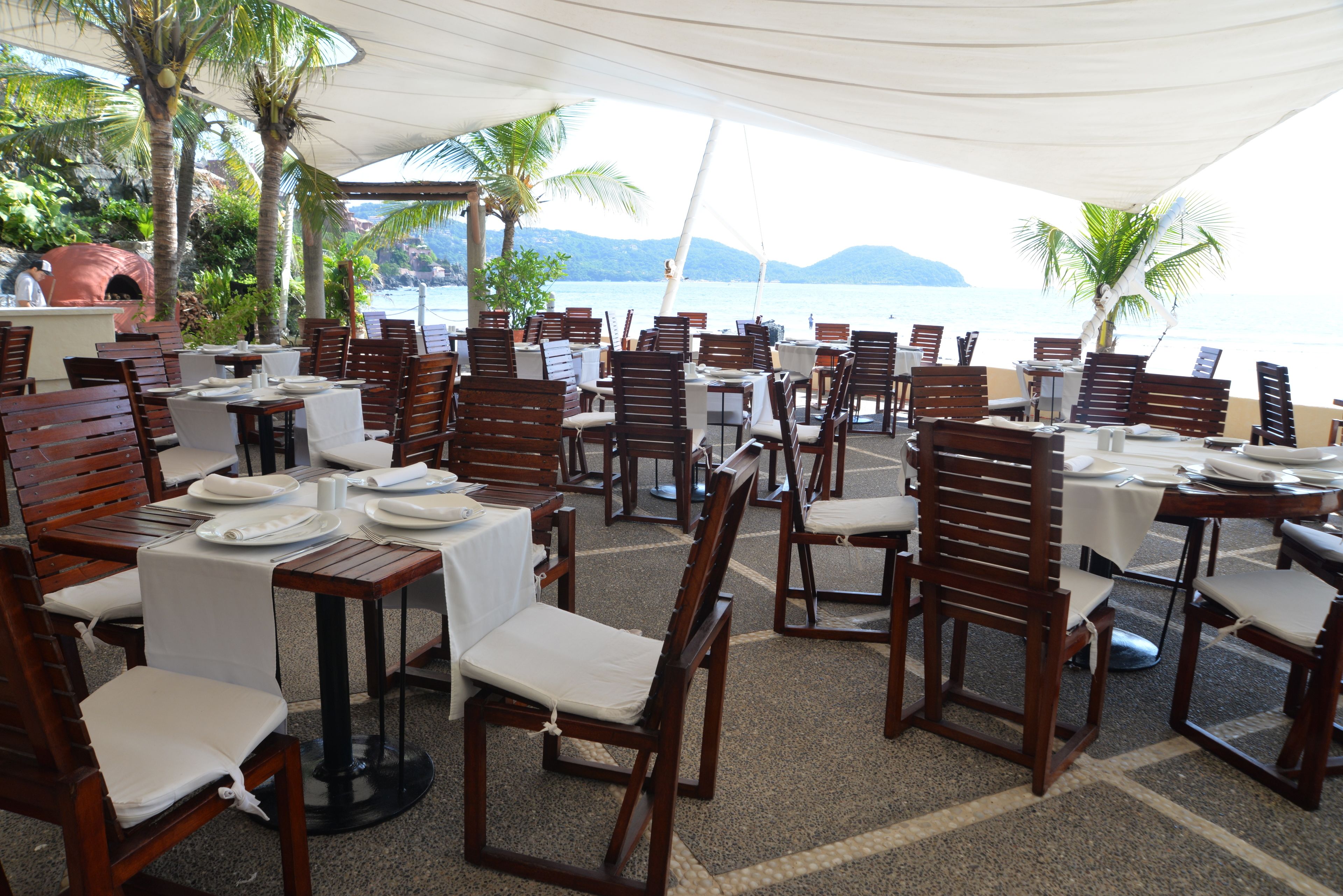 A beachside restaurant with wooden tables and chairs, white tablecloths, and a view of the sea and mountains.
