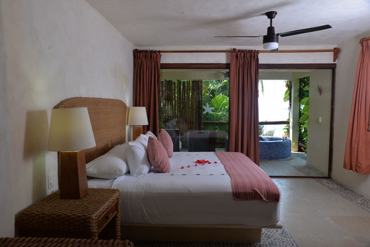 Une chambre d’hôtel avec un lit, deux lampes de chevet, un ventilateur de plafond et une vue sur un jardin, accessible par une porte-fenêtre coulissante.