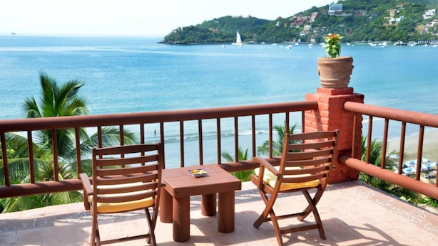 A balcony with two wooden chairs and a small table, overlooking a coastal view with a distant island and a sailboat. - Hotel Aura del Mar (Zihuatanejo)