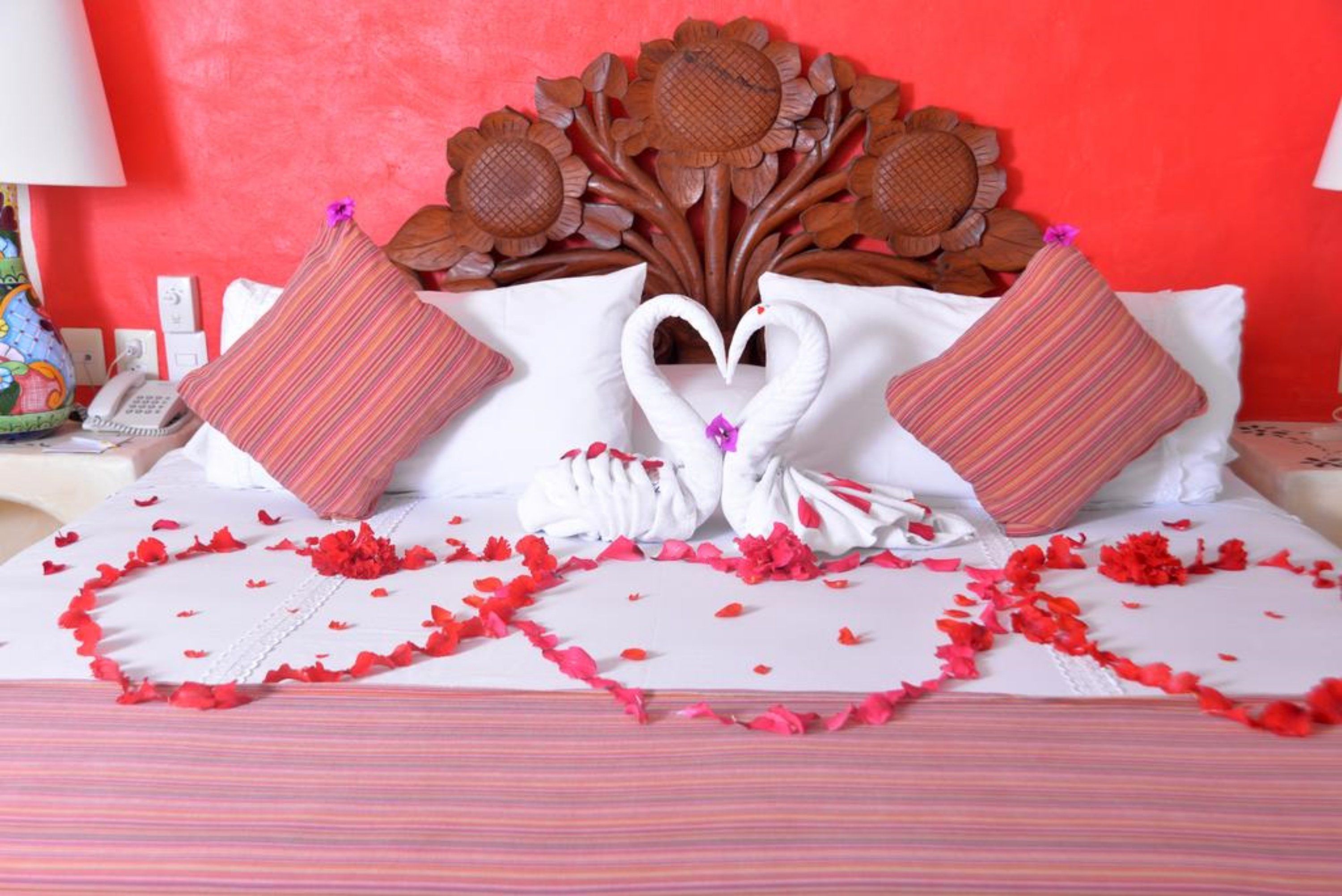 Una cama con disposición de almohadas en forma de corazón, un cabecero decorativo y pétalos de rosa esparcidos.