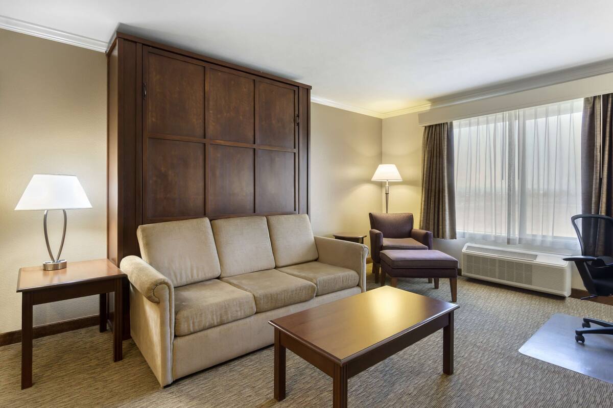 A hotel room with a sofa, armchair, coffee table, and a large wardrobe. There are two lamps and a window with curtains.