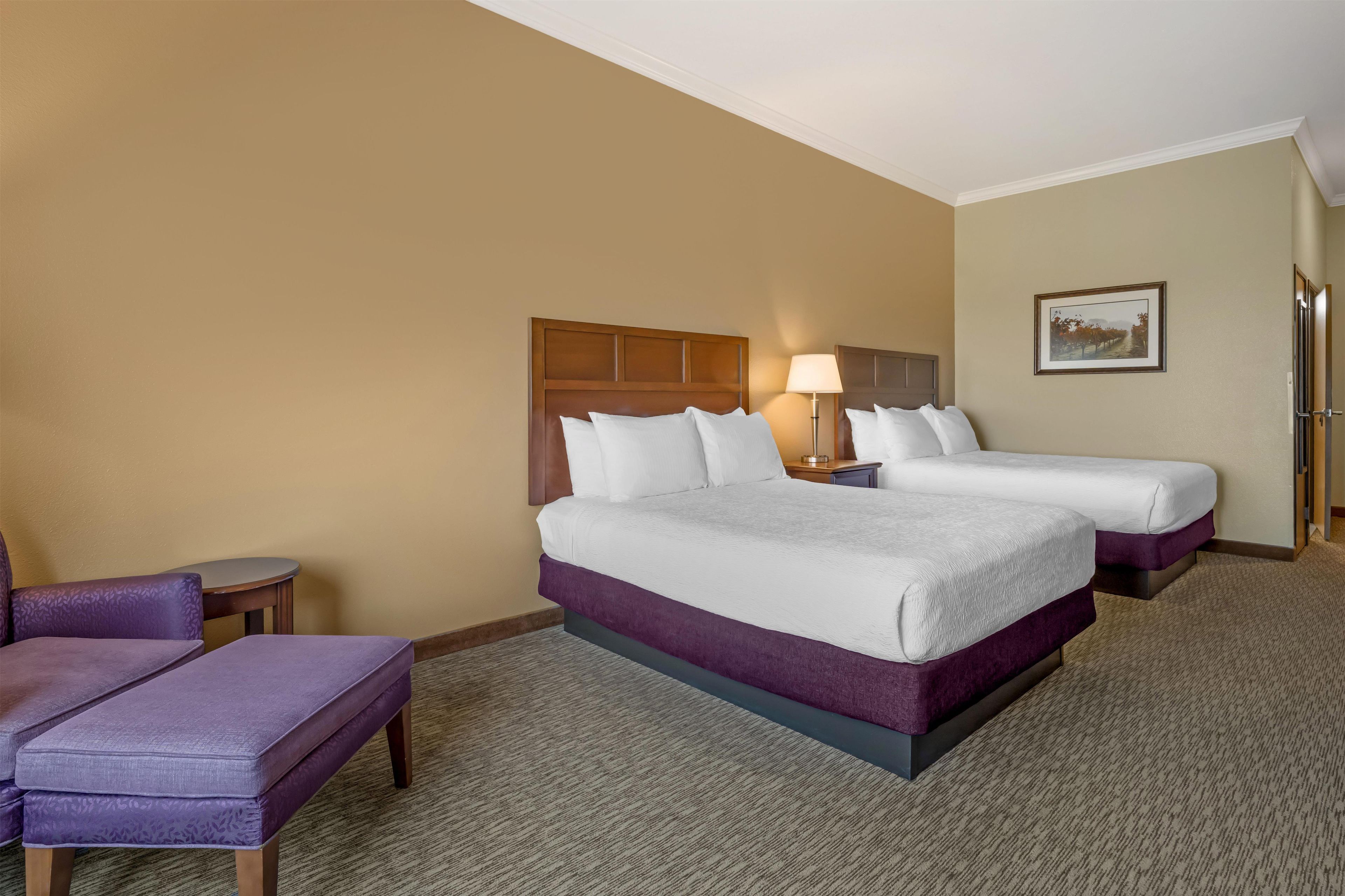 A hotel room with two beds, a small table, a chair, and a lamp.