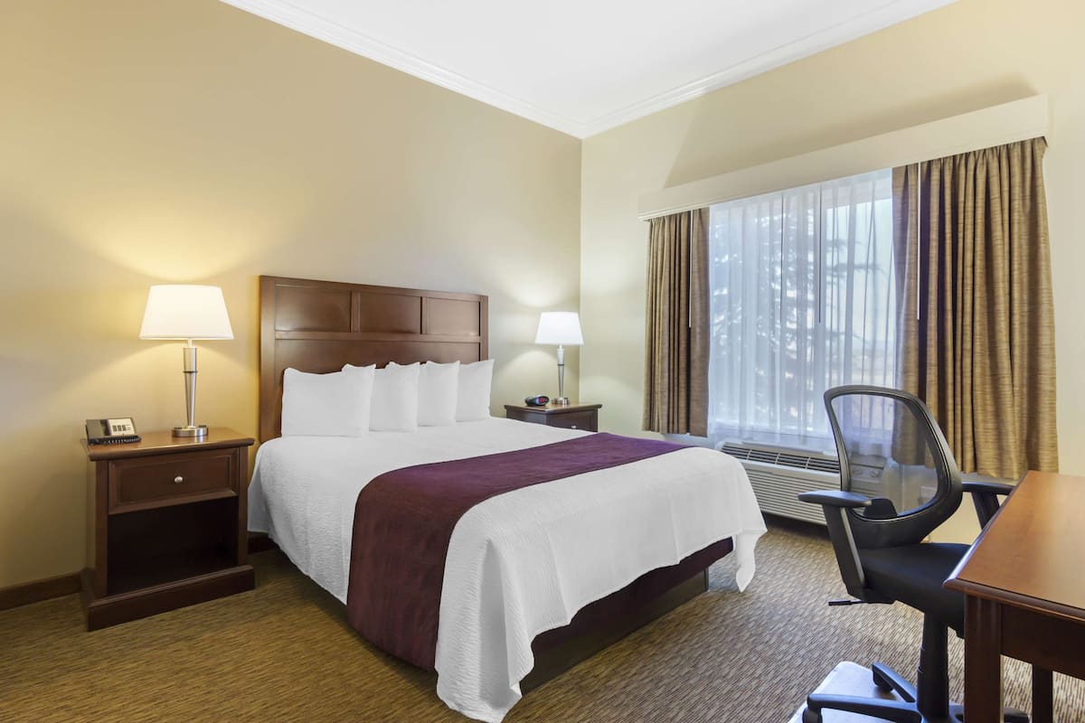 A hotel room with a bed, two bedside tables with lamps, a desk with a chair, and a large window with curtains.