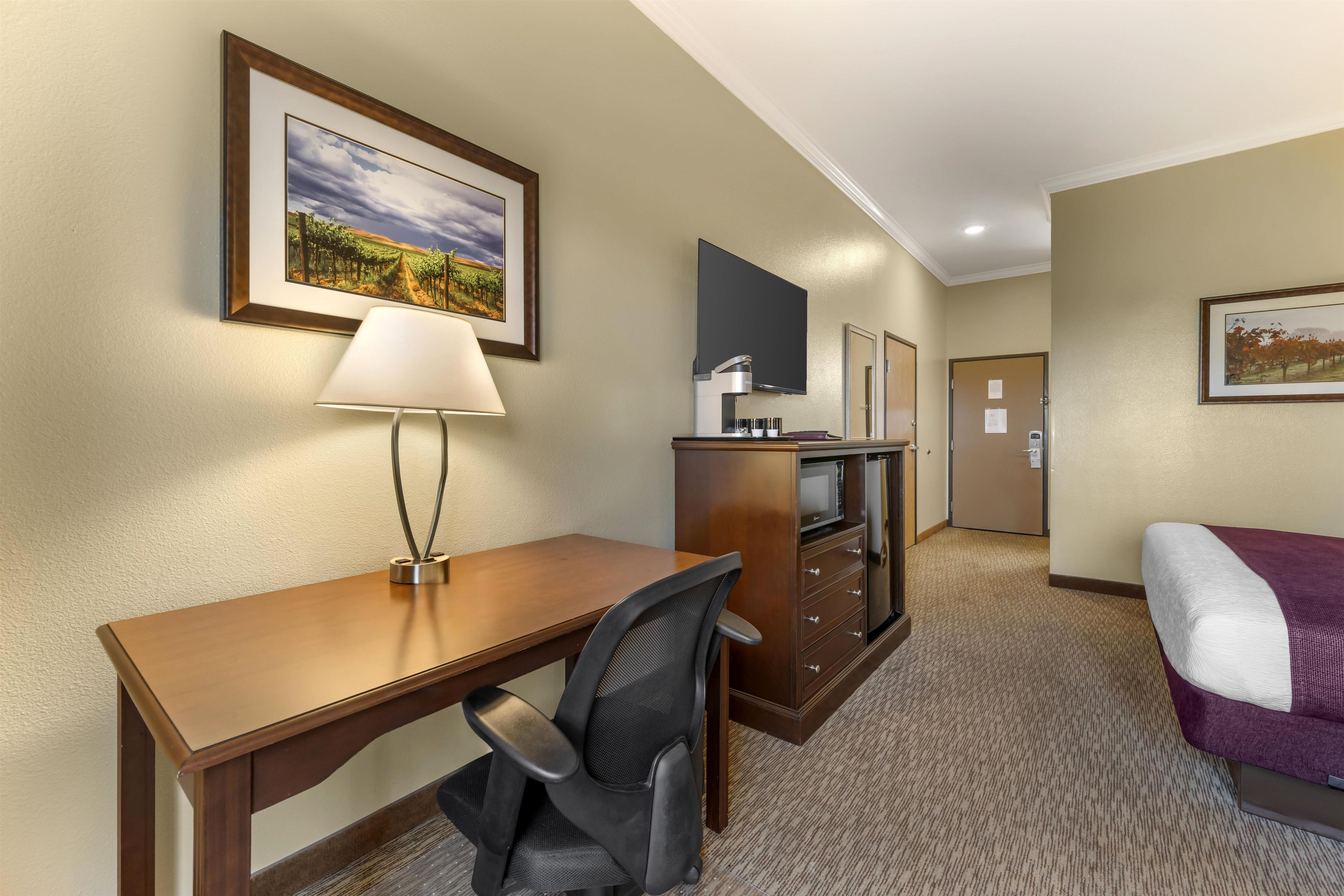 A hotel room with a wooden desk, a chair, a bed with a purple blanket, a television, a minibar, and a door.