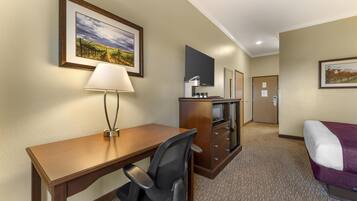 A hotel room with a wooden desk, a chair, a bed with a purple blanket, a television, a minibar, and a door.