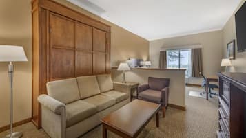 Habitación de hotel con sofá, sillón, una mesita, escritorio con silla, televisor montado en la pared y un ventanal con cortinas.