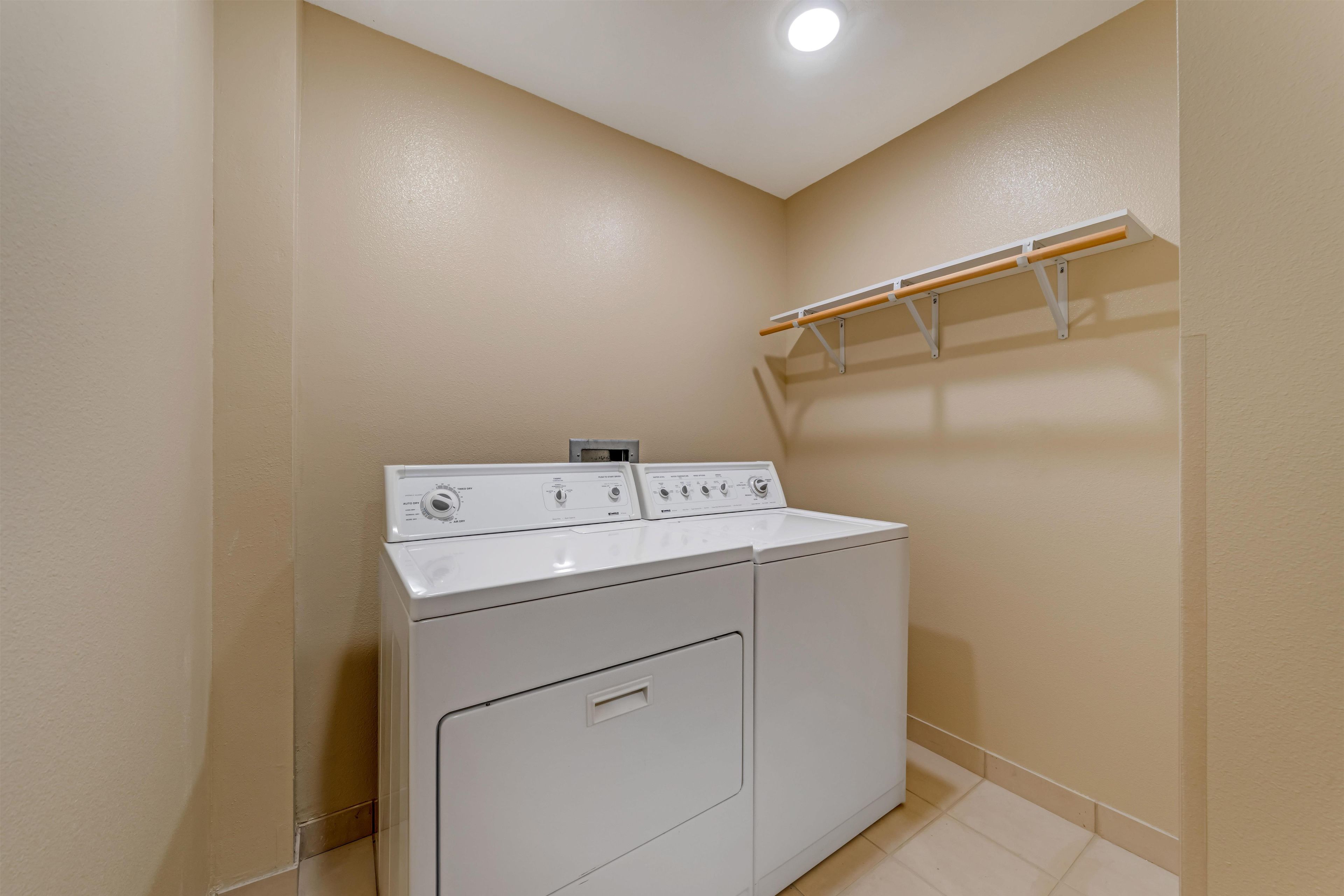 A laundry room with a front-loading washer and dryer, a wooden shelf, and a ceiling light.