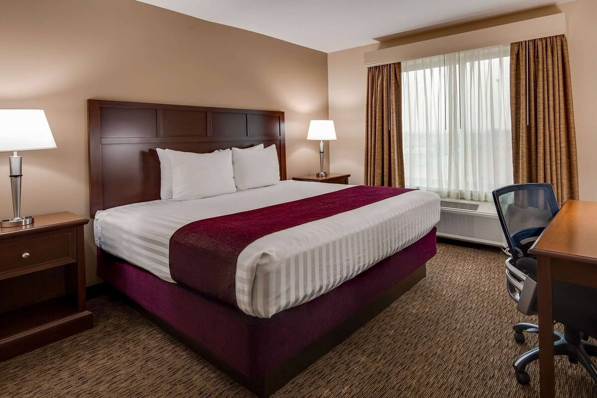 A hotel room with a large bed, two bedside tables with lamps, a desk with a chair, and a window with curtains.