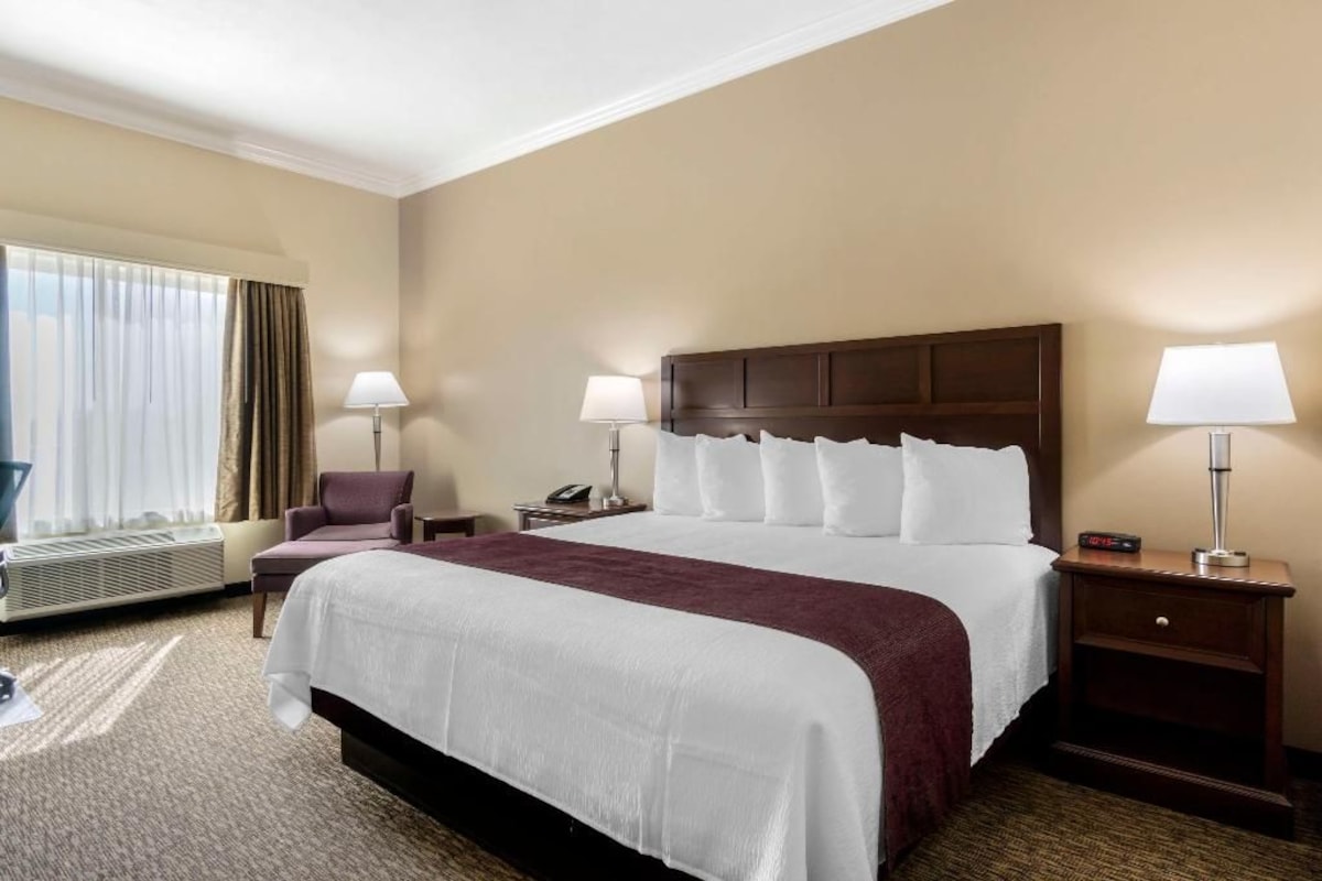 Una habitación de hotel con una cama grande, dos mesitas de noche con lámparas, una silla, un ventanal con cortinas y un equipo de aire acondicionado.