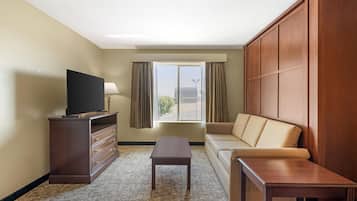 Une chambre d’hôtel avec une télévision posée sur une commode, un canapé, une petite table et une fenêtre avec des rideaux.