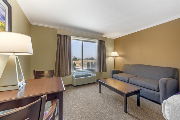 A hotel room with a sofa, a table, a chair, a lamp, a window with curtains, and an air conditioning unit.