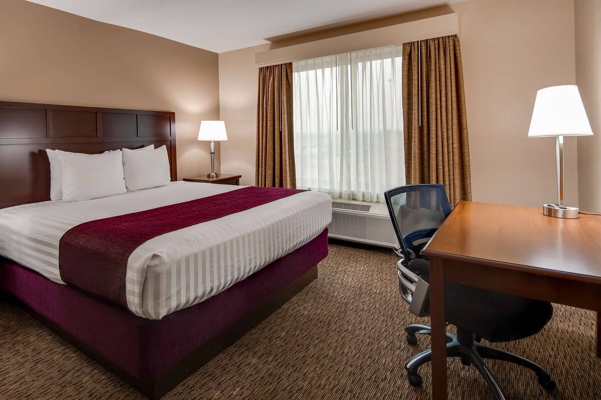 A hotel room with a large bed, two bedside tables with lamps, a desk with a chair, and a window with curtains.