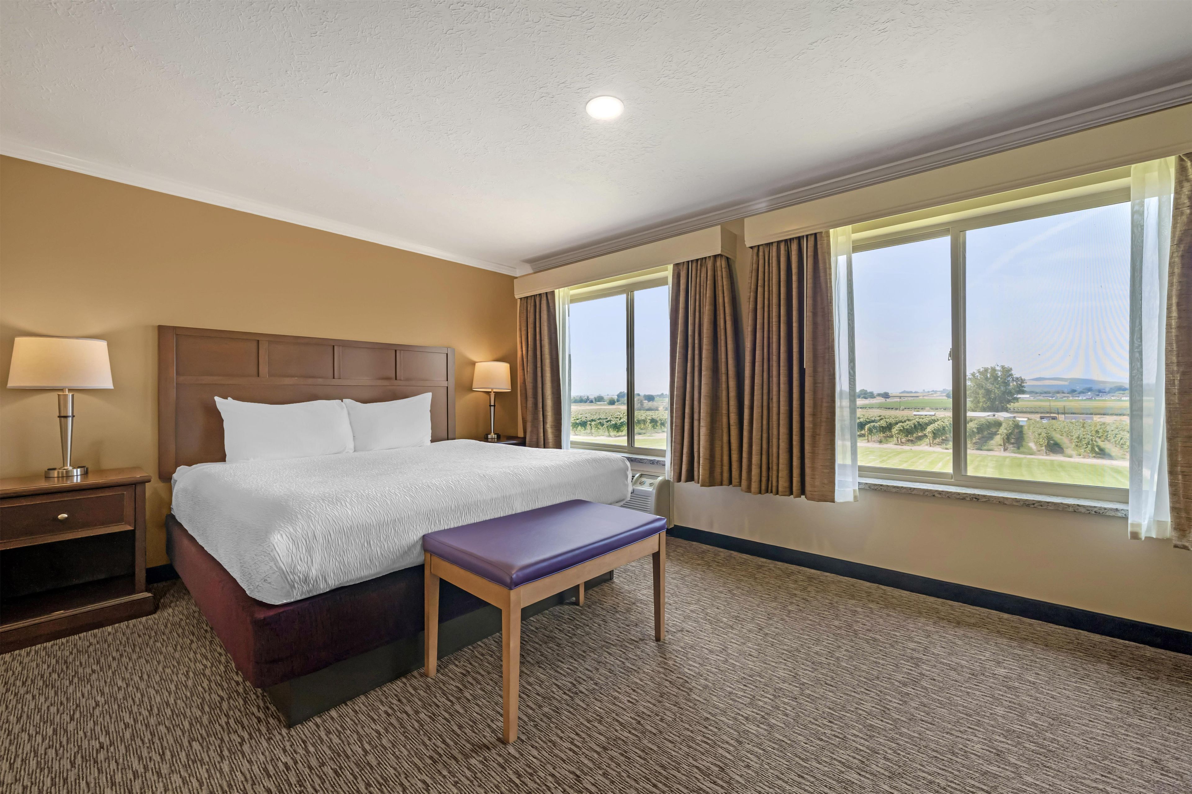 A hotel room with a large bed, two bedside lamps, a bench, and a window with curtains.