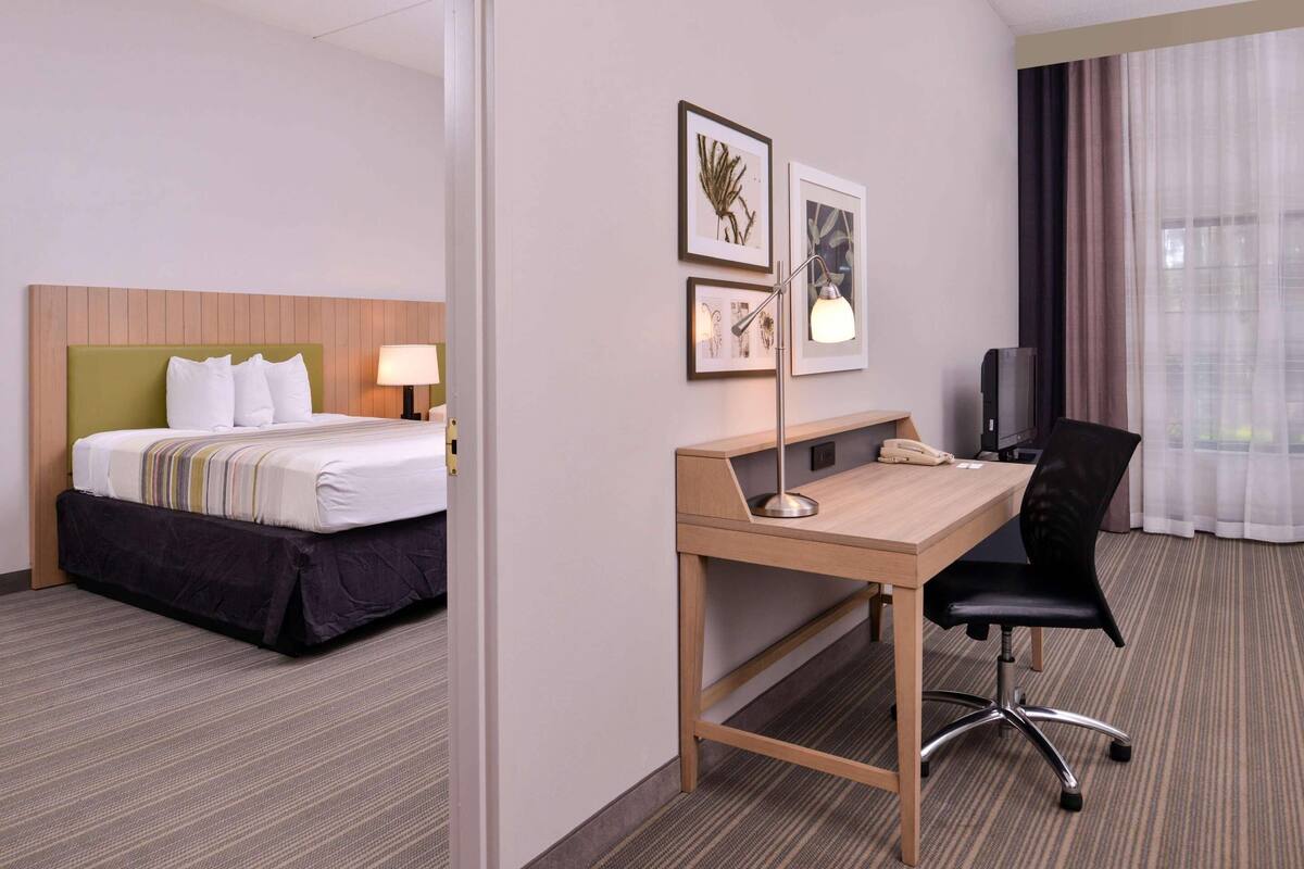 A hotel room with a bed, desk, chair, lamp, and artwork on the wall.