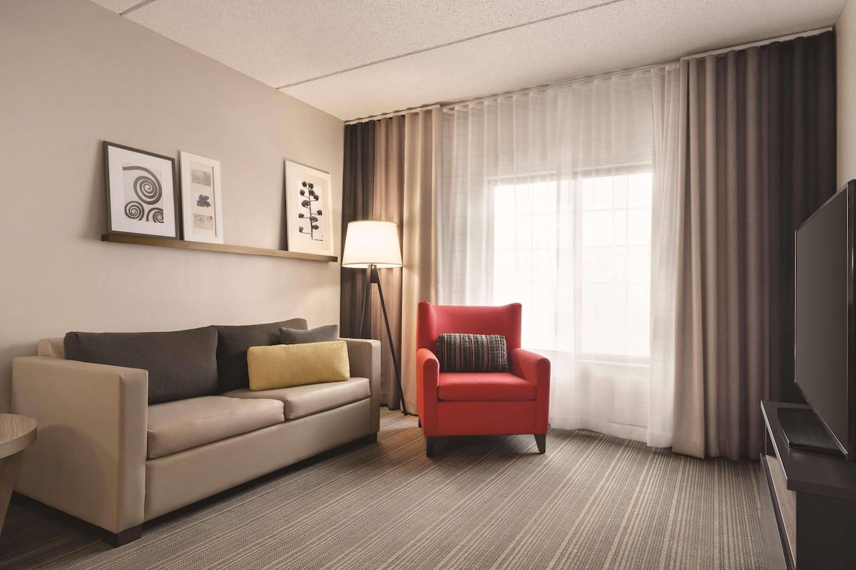 A hotel room with a sofa, armchair, TV, and artwork on the wall.