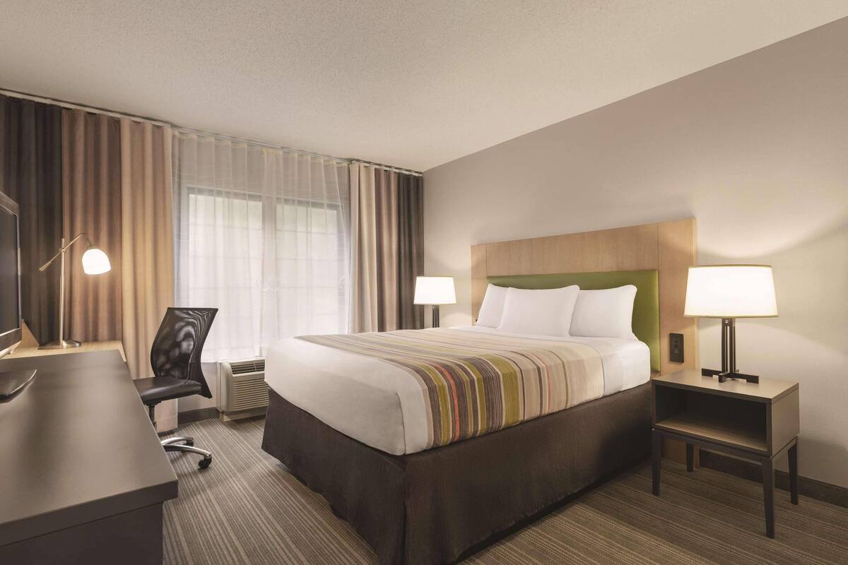 A hotel room with a bed, bedside tables, a desk, a chair, and a large window with curtains.