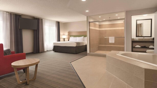 A hotel room with a bed, a red armchair, a small table, a shower area, and a large bathtub. - Country Inn & Suites by Radisson, Raleigh-Durham Airport, NC (Morrisville)
