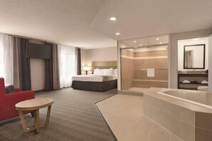 A hotel room with a bed, a red armchair, a small table, a shower area, and a large bathtub.