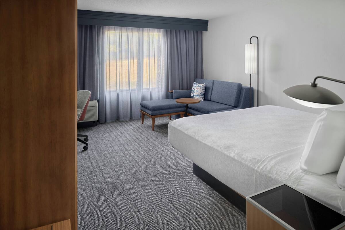 Habitación de hotel con cama, escritorio, silla, sofá, lámpara y ventana con cortinas.