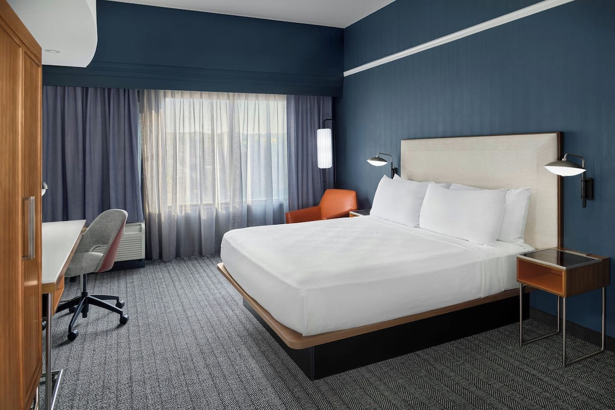 Una habitación de hotel con una cama grande, un escritorio con silla, una mesita de noche y una ventana con cortinas.