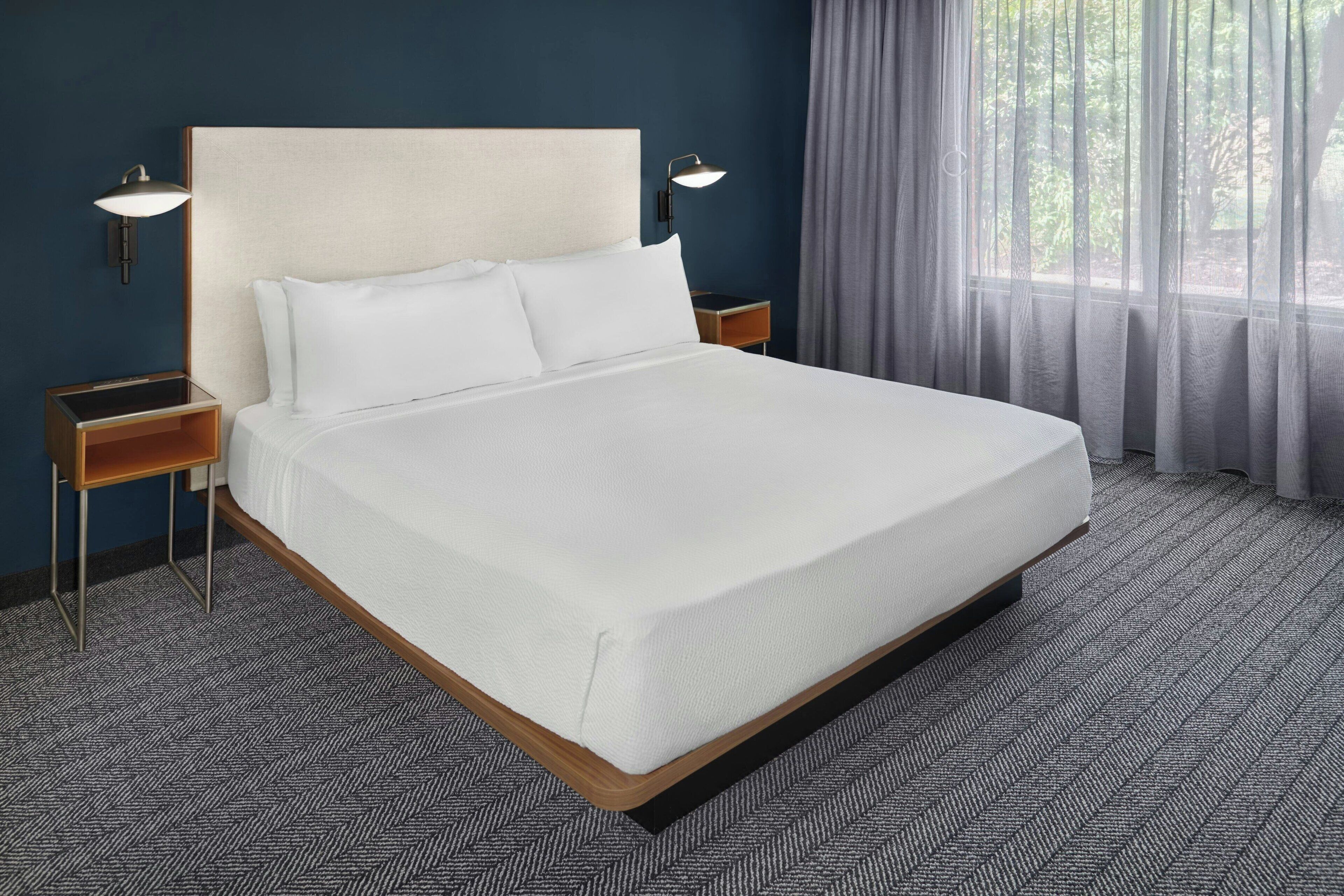 A neatly made bed with white linens in a hotel room. There are two bedside tables with lamps, a window with sheer curtains, and a herringbone-patterned carpet.